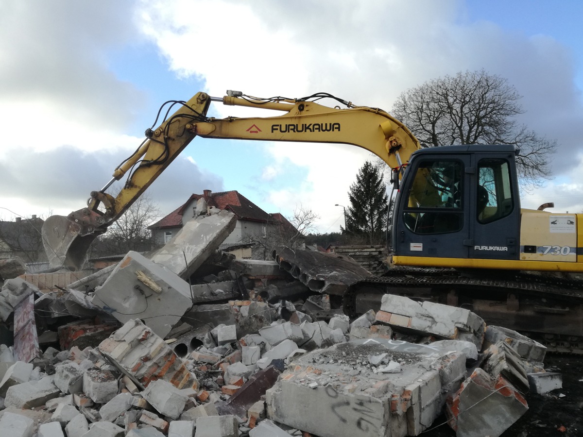 Żółto-szara koparka marki Furukawa 730 na gruzowisku z cegieł i betonu, w tle dom z czerwonym dachem i pochmurne niebo.