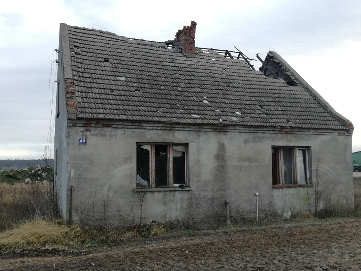 Zrujnowany, jednopiętrowy budynek z częściowo zawalonym dachem i wybitymi oknami, widoczny numer domu '46' na elewacji, w tle sterta gruzu.