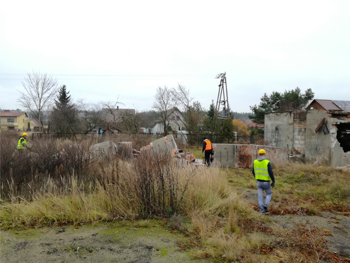 Trzech pracowników w odblaskowych kamizelkach i kaskach na terenie po zburzonym budynku z gruzem, widoczne fragmenty ścian i fundamentów, w tle zabudowa jednorodzinna i słup energetyczny.