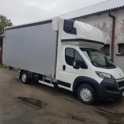 Biały samochód dostawczy Peugeot z szarą plandeką na mokrym podłożu, zaparkowany przed budynkiem z szarymi ścianami i małymi oknami.