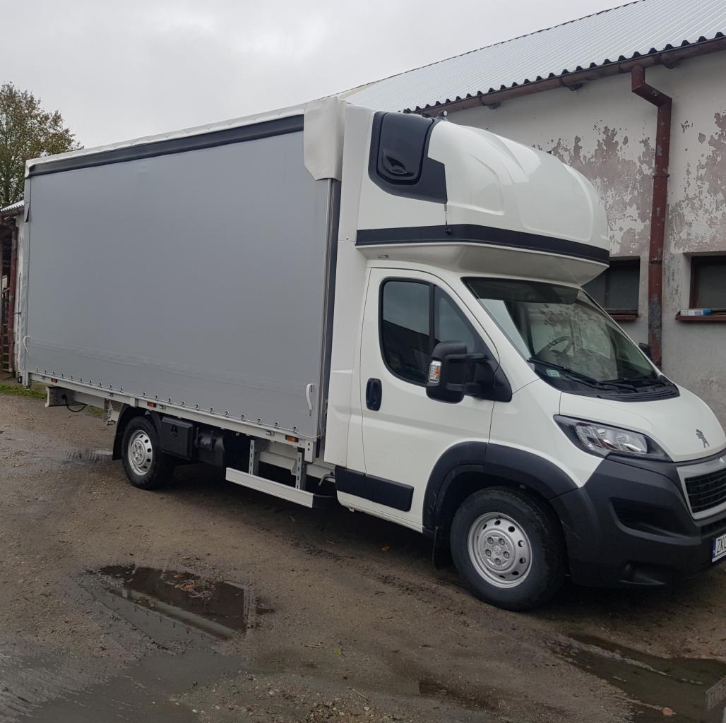 Biały samochód dostawczy Peugeot z szarą plandeką na mokrym podłożu, zaparkowany przed budynkiem z szarymi ścianami i małymi oknami.