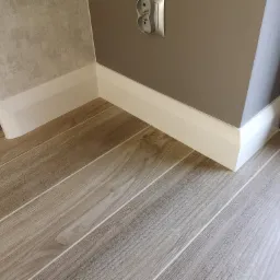 Ułożona podłoga z płytek imitujących drewno, białe listwy przypodłogowe przy szarej ścianie z gniazdkiem elektrycznym.