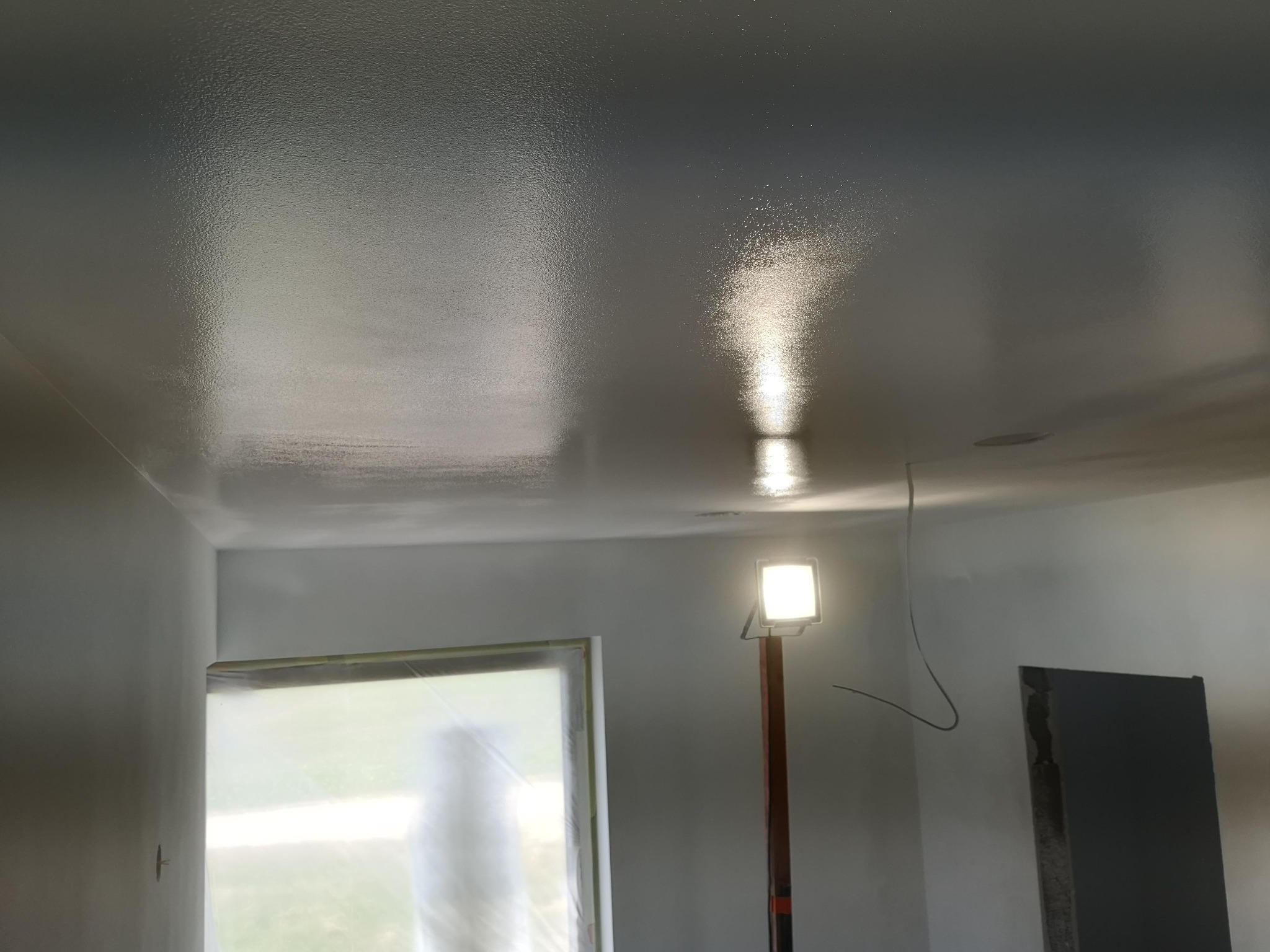 Świeżo pomalowany, błyszczący sufit w pokoju, oświetlony lampą budowlaną, z widocznym zabezpieczonym oknem i fragmentem ściany z niedokończonym wykończeniem.
