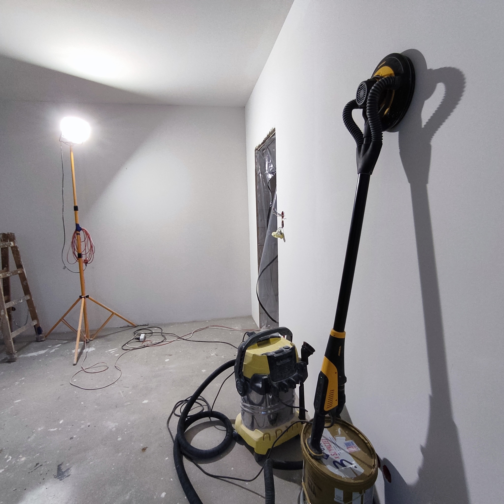Wnętrze pokoju w trakcie prac wykończeniowych: szlifierka do gładzi na wiadrze po farbie, odkurzacz przemysłowy, lampa budowlana na trójnogu i drabina malarska w tle.