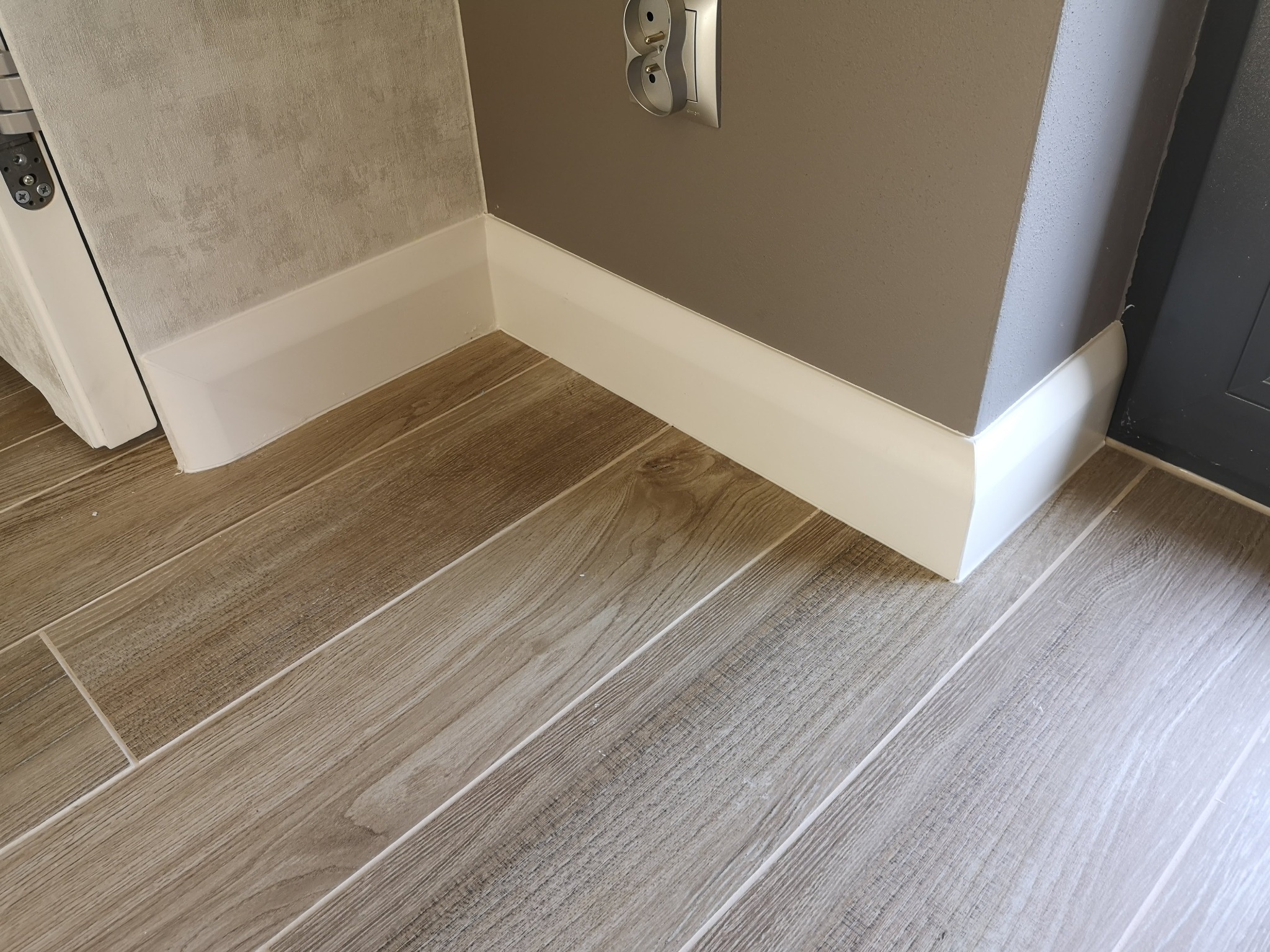 Ułożona podłoga z płytek imitujących drewno, białe listwy przypodłogowe przy szarej ścianie z gniazdkiem elektrycznym.