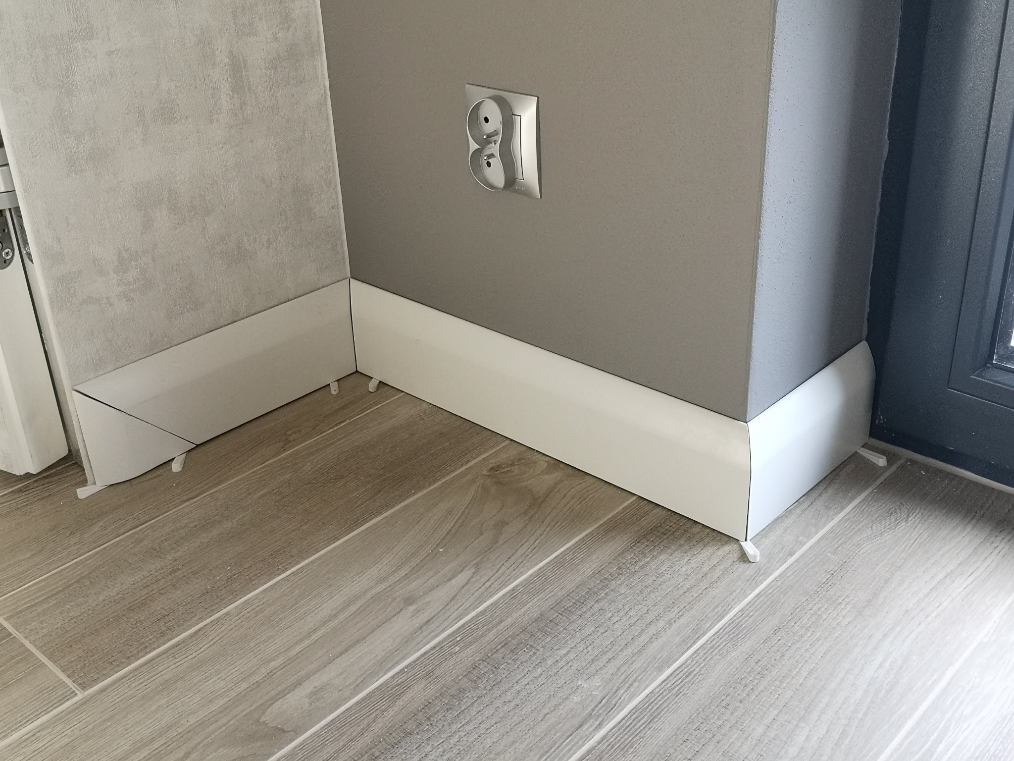 Szczegółowe ujęcie nowo zamontowanych, białych listew przypodłogowych z widocznymi, plastikowymi klinami dystansowymi, na tle szarej ściany z gniazdkiem elektrycznym i podłogi z paneli imitujących...