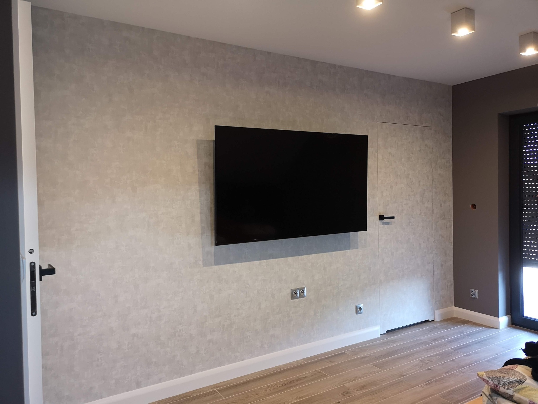 Jasnoszara ściana z efektem betonu, na której wisi czarny telewizor, widoczne gniazdka elektryczne i ukryte drzwi z czarną klamką, podłoga wyłożona panelami imitującymi drewno.