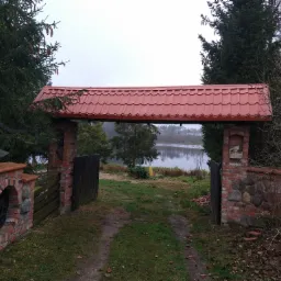 Brama z czerwonym dachem z dachówki, zbudowana z cegły i kamienia, prowadząca na brzeg jeziora w pochmurny dzień.