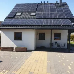 Dom jednorodzinny z panelami słonecznymi na dachu i dwoma oknami dachowymi, widok z zewnątrz, kostka brukowa w dwóch odcieniach na podjeździe.