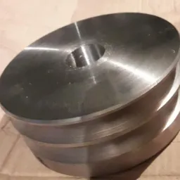 Szara, metalowa część z otworem na środku i widocznymi śladami obróbki, leżąca na kartonie.