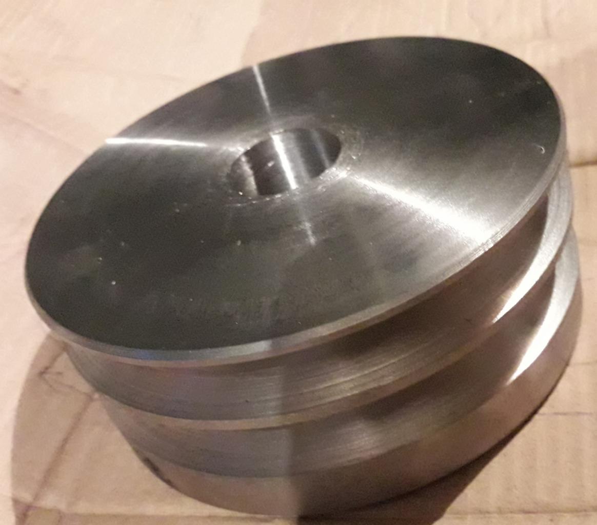 Szara, metalowa część z otworem na środku i widocznymi śladami obróbki, leżąca na kartonie.