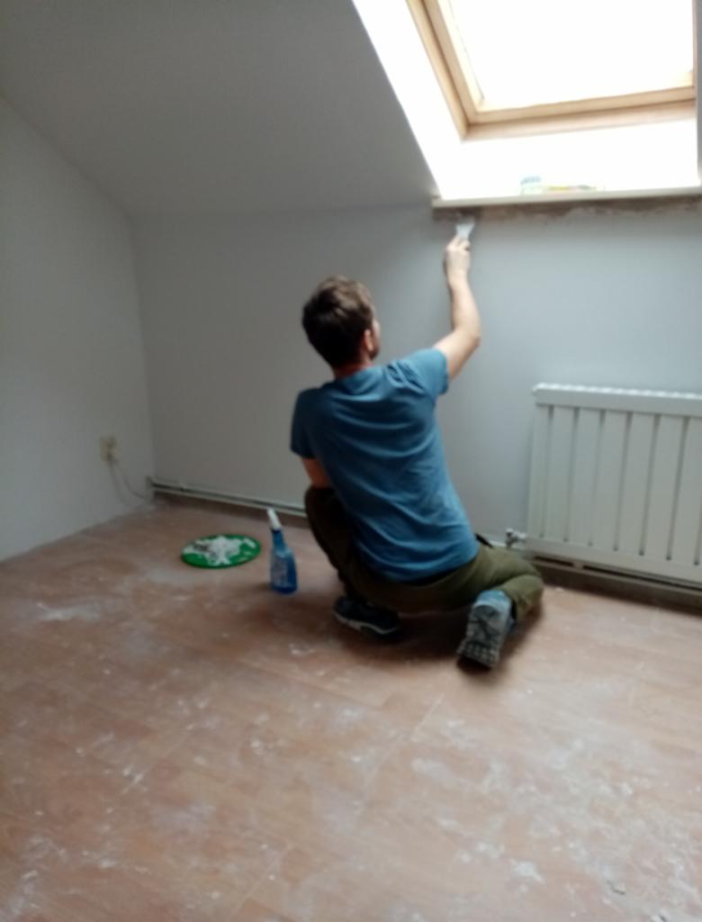 Malowanie pokoju na poddaszu: mężczyzna maluje ścianę pod oknem dachowym, widoczne narzędzia i ślady farby na podłodze. Grzejnik i gniazdko w tle.