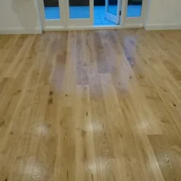 Jasna, drewniana podłoga ułożona w salonie z białymi listwami przypodłogowymi i przeszklonymi drzwiami balkonowymi w tle.