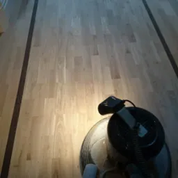 Maszyna do polerowania parkietu z widocznymi ciemnymi bordiurami przy ścianach, oświetlona punktowo, w tle fragment szafki.