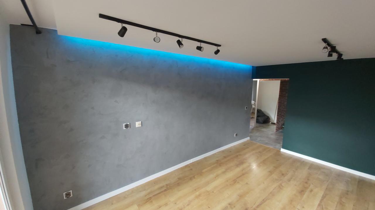 Szara ściana z efektem dekoracyjnym, podświetlona niebieskim światłem LED, z zamontowanymi czarnymi reflektorami sufitowymi i drewnianą podłogą.