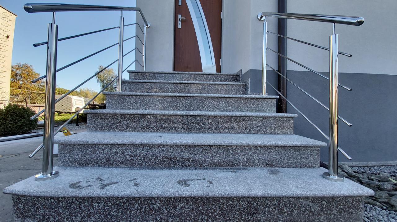 Granitowe schody zewnętrzne z nowoczesną, stalową balustradą, prowadzące do wejścia budynku z drewnianymi drzwiami.