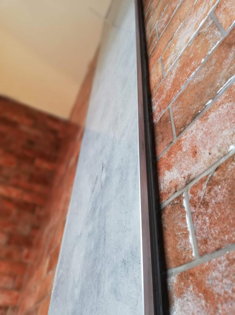 Szara płyta imitująca beton, zamontowana na ścianie z czerwonej cegły, widoczna metalowa listwa wykończeniowa.