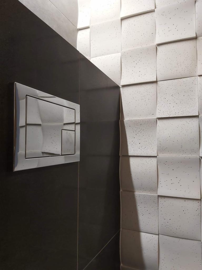 Nowoczesna łazienka z czarną, błyszczącą ścianą i chromowanym przyciskiem spłuczki oraz trójwymiarową, białą ścianą z małymi, ciemnymi plamkami.