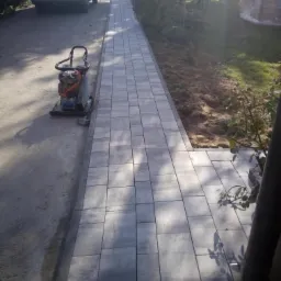 Świeżo ułożona, szara kostka brukowa tworząca ścieżkę w ogrodzie, z ubijarką wibracyjną obok i samochodem w tle.