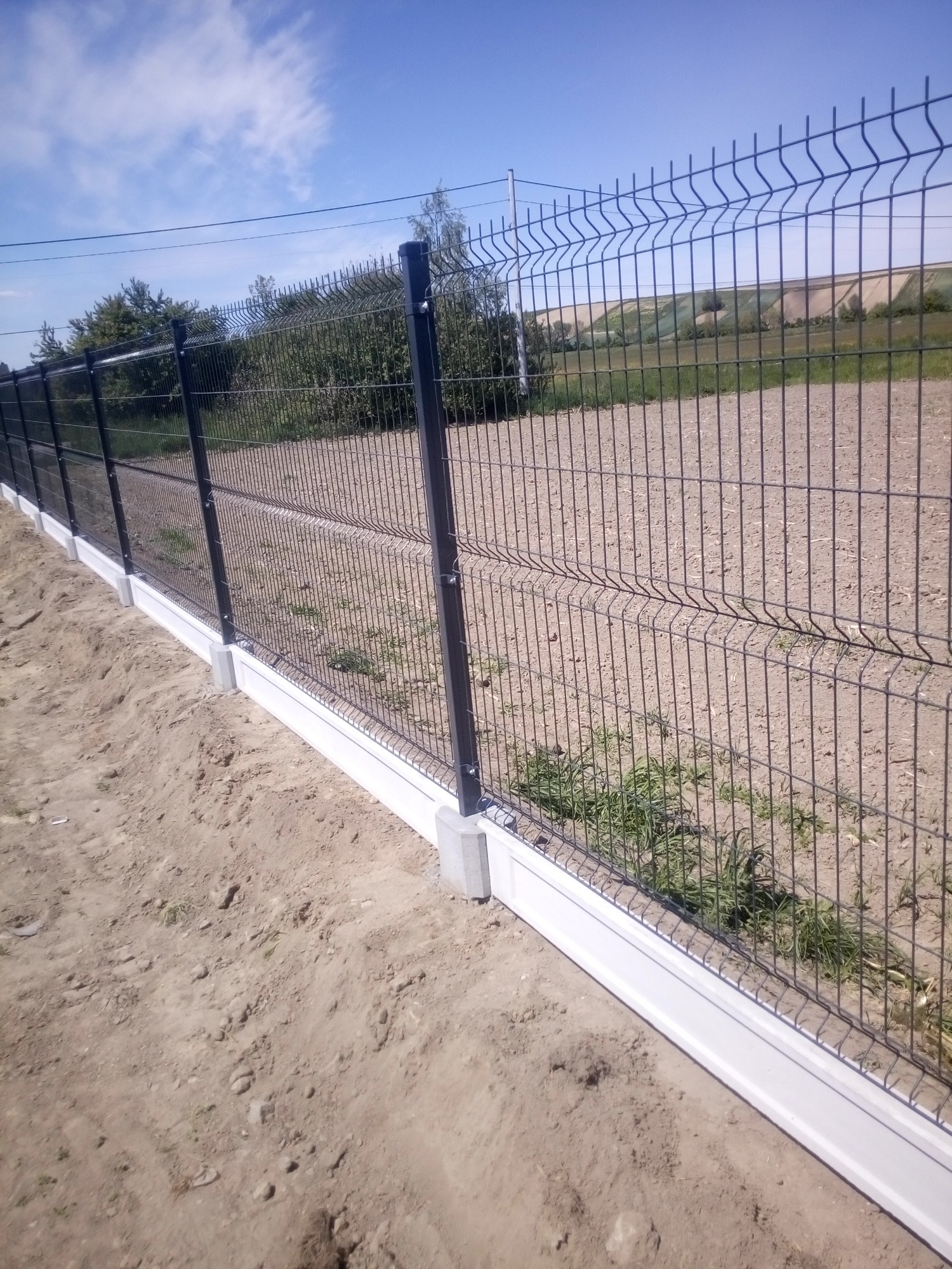 Nowoczesne ogrodzenie panelowe z podmurówką prefabrykowaną, widok z bliska na grunt i fragment pola w tle.