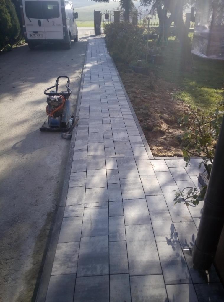 Świeżo ułożona, szara kostka brukowa tworząca ścieżkę w ogrodzie, z ubijarką wibracyjną obok i samochodem w tle.