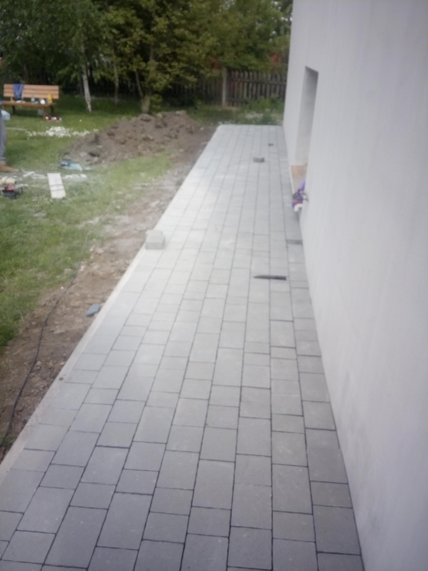 Świeżo ułożona szara kostka brukowa przy białej ścianie budynku, widoczny fragment trawnika z narzędziami i pryzmą ziemi w tle.