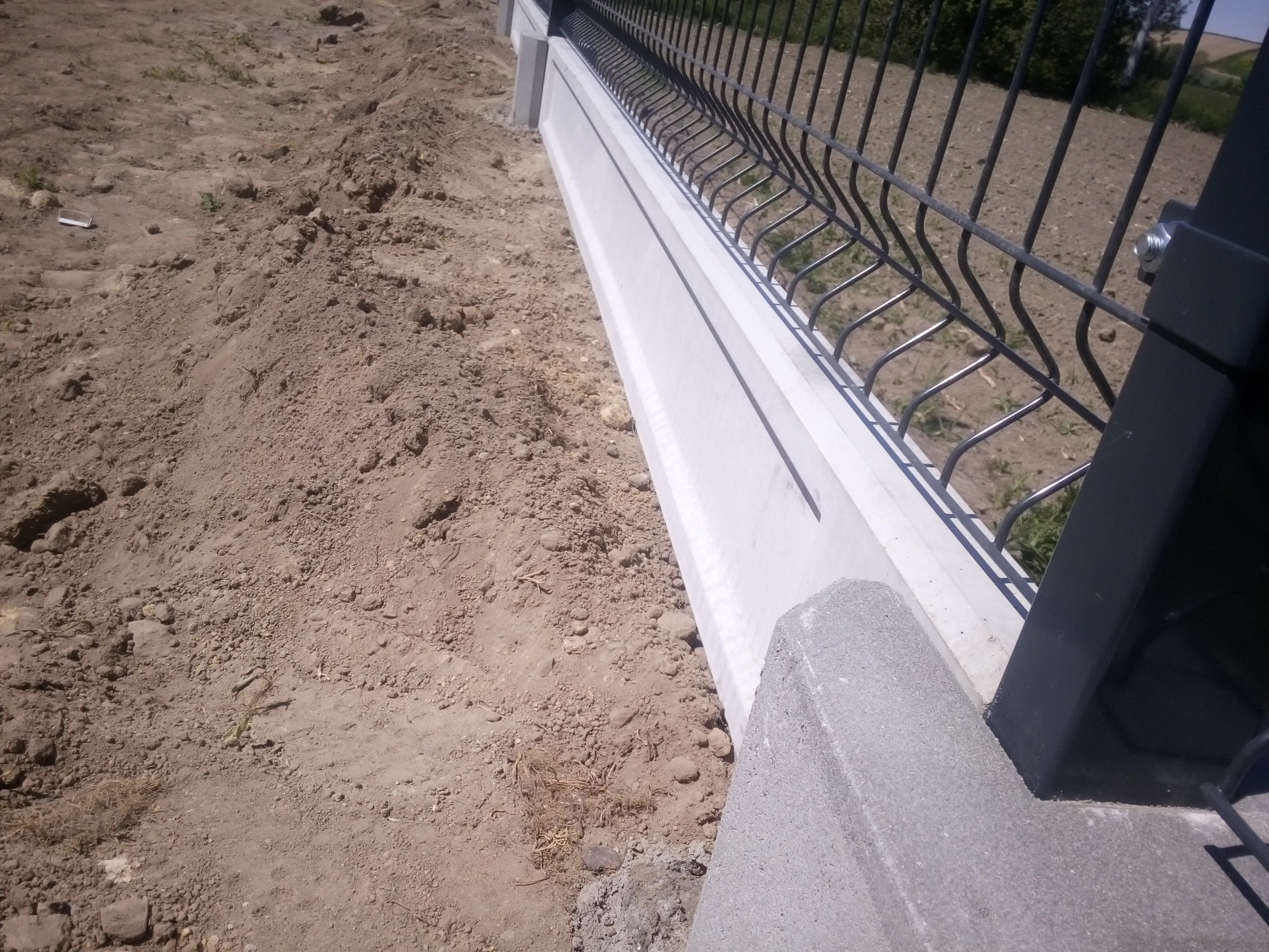 Fragment nowo postawionego ogrodzenia panelowego z podmurówką prefabrykowaną na nierównym, gliniastym gruncie. Widoczne metalowe słupki i siatka panelowa w kolorze grafitowym.