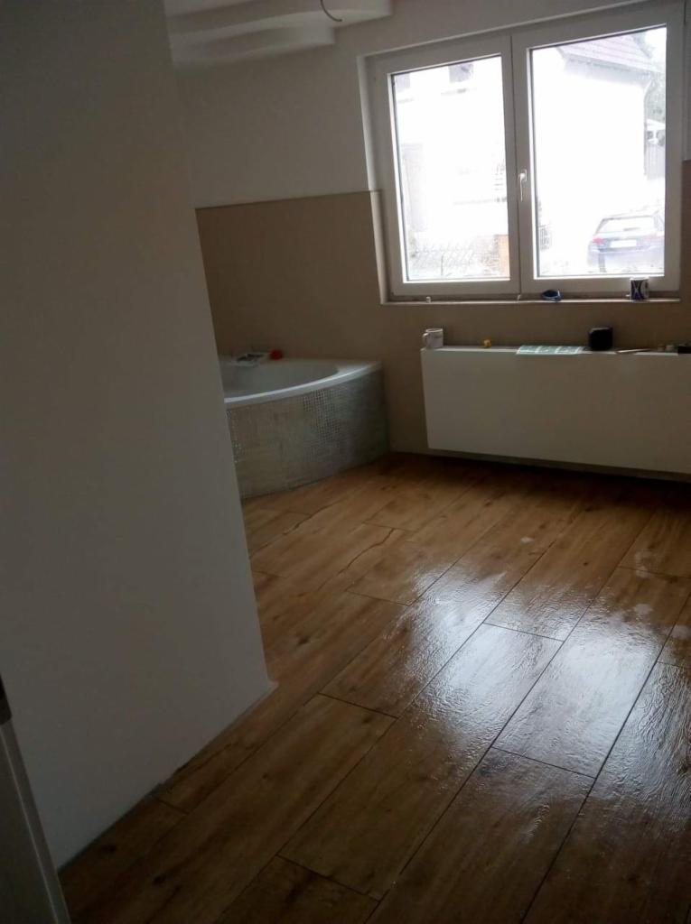 Wykończona łazienka z drewnopodobną podłogą, widoczna wanna z mozaiką i grzejnik pod oknem