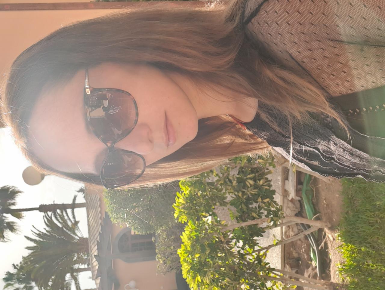 Portret kobiety w okularach przeciwsłonecznych na tle egzotycznej roślinności i budynku z łukowatym oknem.