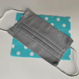 Szara, plisowana maseczka ochronna z elastycznymi gumkami, leżąca na kawałku turkusowej tkaniny w białe grochy, na białym tle.