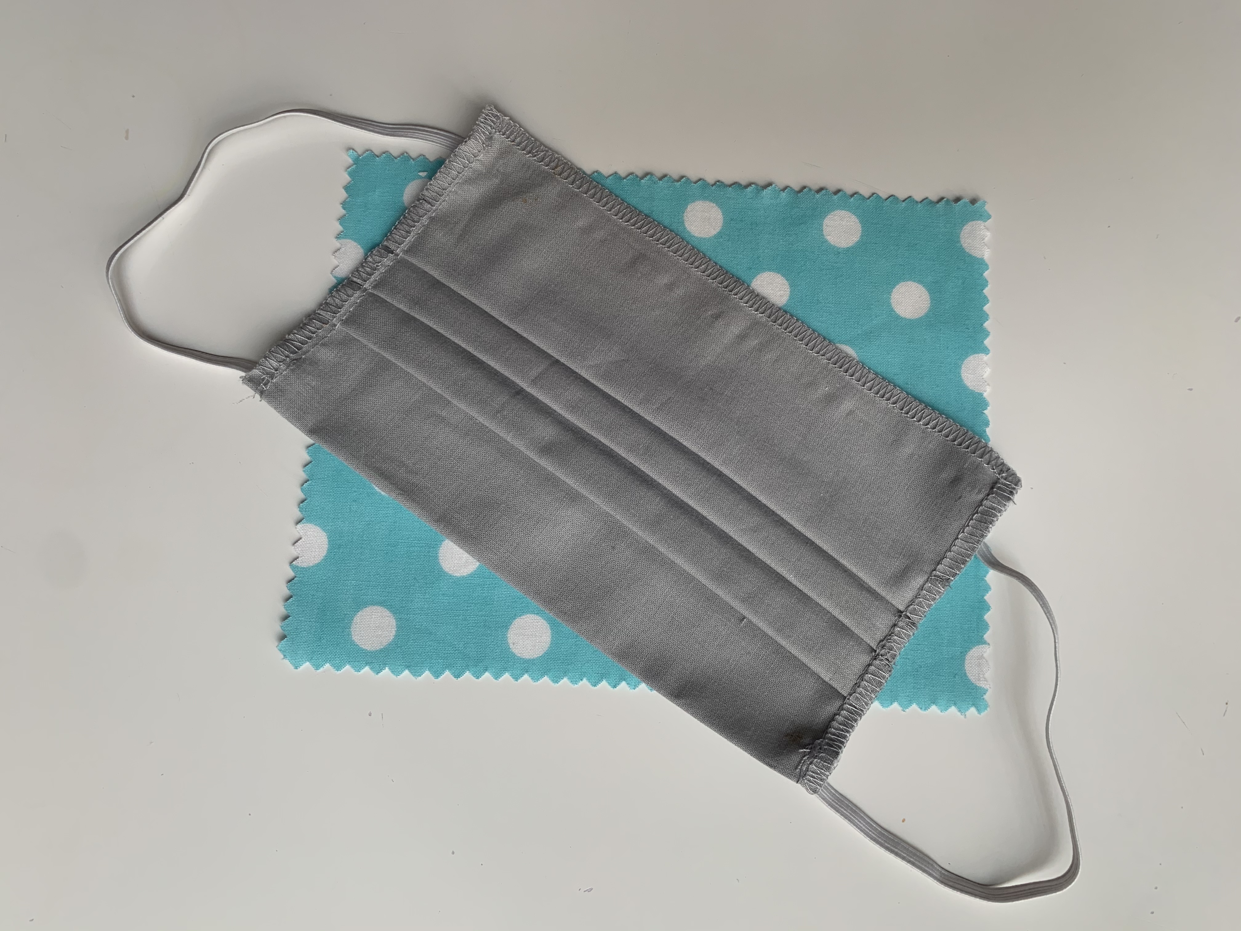 Szara, plisowana maseczka ochronna z elastycznymi gumkami, leżąca na kawałku turkusowej tkaniny w białe grochy, na białym tle.