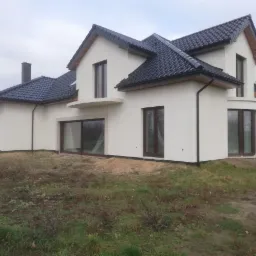 Wykończony dom jednorodzinny z białą elewacją, ciemną dachówką i brązowymi oknami, widok z zewnątrz na niezagospodarowanej działce.