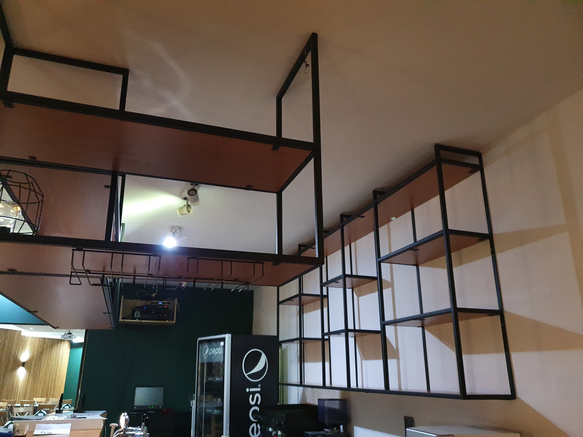 Metalowa konstrukcja z drewnianymi półkami, prawdopodobnie regał barowy, z uchwytami na kieliszki, widziana z dołu, w tle bar z lodówką Pepsi.