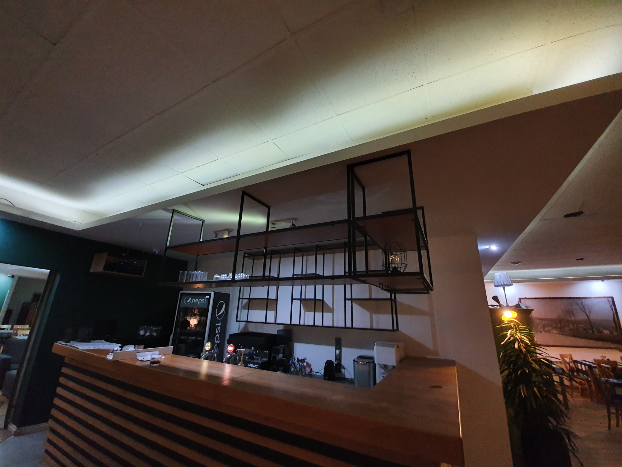 Widok na bar z drewnianym blatem, za którym znajduje się ekspres do kawy, lodówka Pepsi i metalowe regały z oświetleniem, w tle widoczne stoliki restauracyjne.