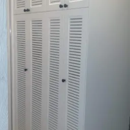 Biała szafa wnękowa z lamelkowymi drzwiami i czarnymi gałkami, umieszczona w rogu pomieszczenia z drewnianą podłogą.