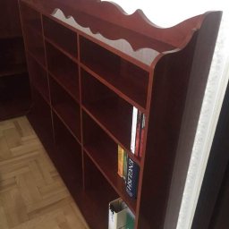 Kamil Matuszewski - Regał z ciemnego drewna z dekoracyjnym wykończeniem na górze, częściowo wypełniony książkami i segregatorami, ustawiony na parkiecie.