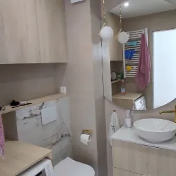 Jasna łazienka z zabudową meblową z drewnopodobnej płyty, okrągłym lustrem i wiszącą lampą z kulistymi kloszami; pralka wkomponowana w zabudowę.