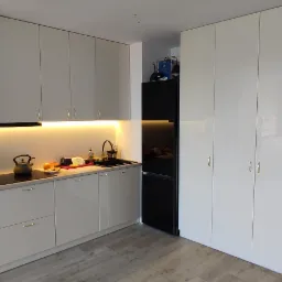 Jasna kuchnia z błyszczącymi frontami, podświetleniem LED podszafkowym i czarną lodówką w zabudowie, obok wysoka szafa wnękowa z czterema drzwiami i widoczny fragment drewnianych drzwi.