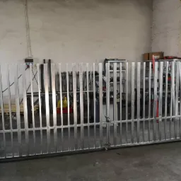 Surowa metalowa brama przemysłowa w trakcie produkcji, widoczne spawy i niedoskonałości obróbki, warsztatowe tło z półkami i narzędziami.