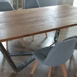 Stół z blatem z drewna o wyraźnym usłojeniu i metalową podstawą w kształcie litery V, otoczony szarymi krzesłami w minimalistycznym stylu.