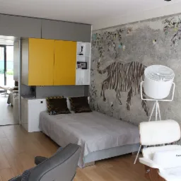 Nowoczesna aranżacja małej sypialni z żółtymi szafkami wiszącymi, łóżkiem, fotelami i tapetą z motywem tygrysa i kwiatów.