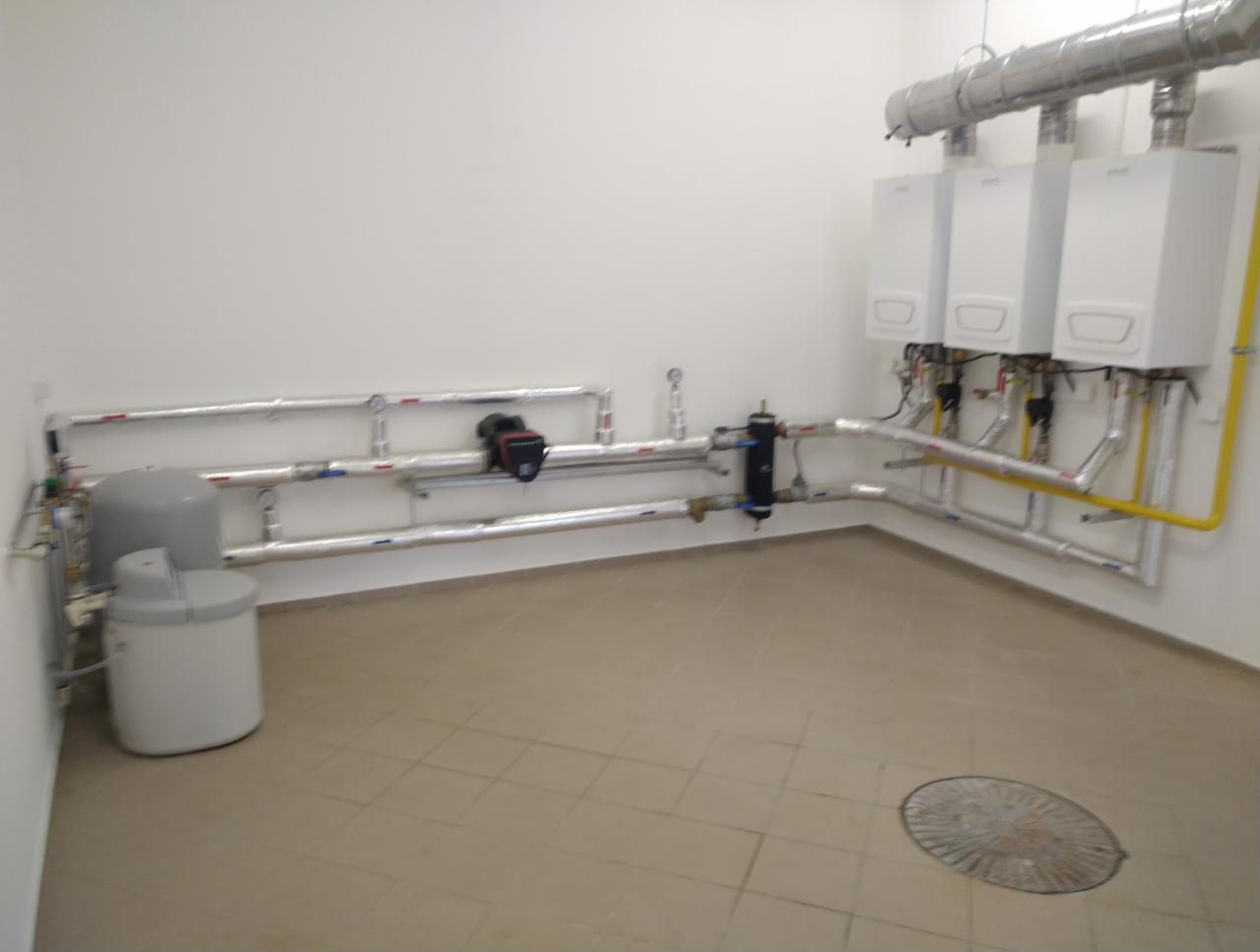Trzy białe kotły grzewcze wiszące na ścianie, połączone rurami z elementami instalacji w pomieszczeniu z płytkami na podłodze i szarym zbiornikiem.