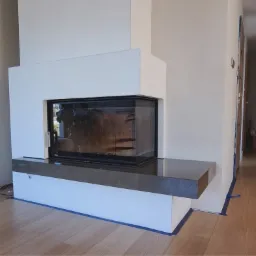 Nowoczesny kominek narożny z czarną ramą i szarą półką, wbudowany w białą ścianę, widoczne taśmy malarskie na podłodze.