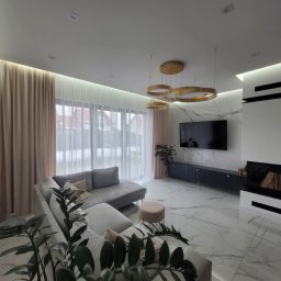 KOMINKI DLA WYMAGAJĄCYCH Wojciech Kaszkowiak - Elegancki salon z kominkiem, marmurową ścianą i podłogą. Nowoczesne oświetlenie, szara sofa i zasłony tworzą spójną całość. Widok na zewnątrz przez okno.