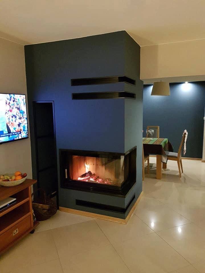 Nowoczesny kominek narożny z czarną obudową i płomieniami, wkomponowany w ciemnoniebieską ścianę z dekoracyjnymi wnękami, w jasnym wnętrzu z telewizorem i stołem.