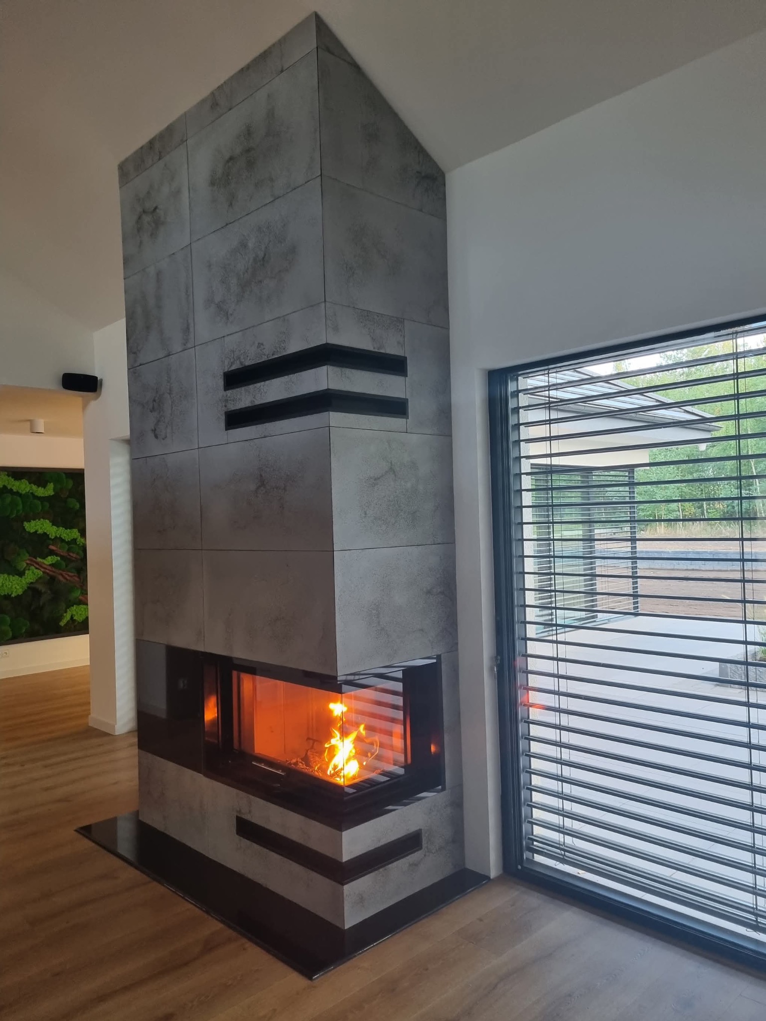 Nowoczesny kominek obłożony szarymi płytami z czarnymi akcentami, płomienie widoczne przez szybę, obok okno z żaluzjami. Minimalistyczny design.