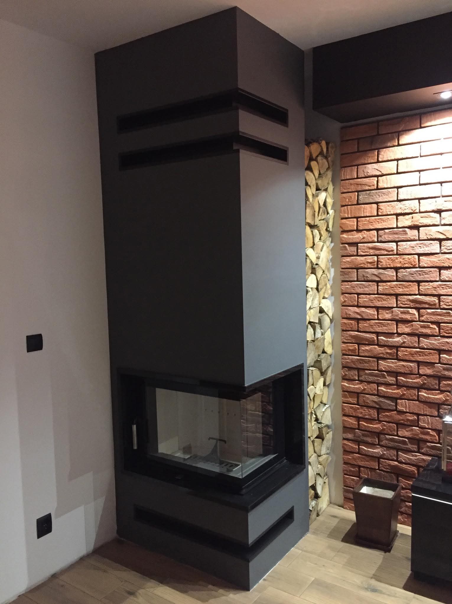 Nowoczesny kominek narożny z obudową w odcieniach szarości, z dekoracyjną ścianą z cegły i ułożonym drewnem obok. Minimalistyczny design, oświetlenie punktowe.