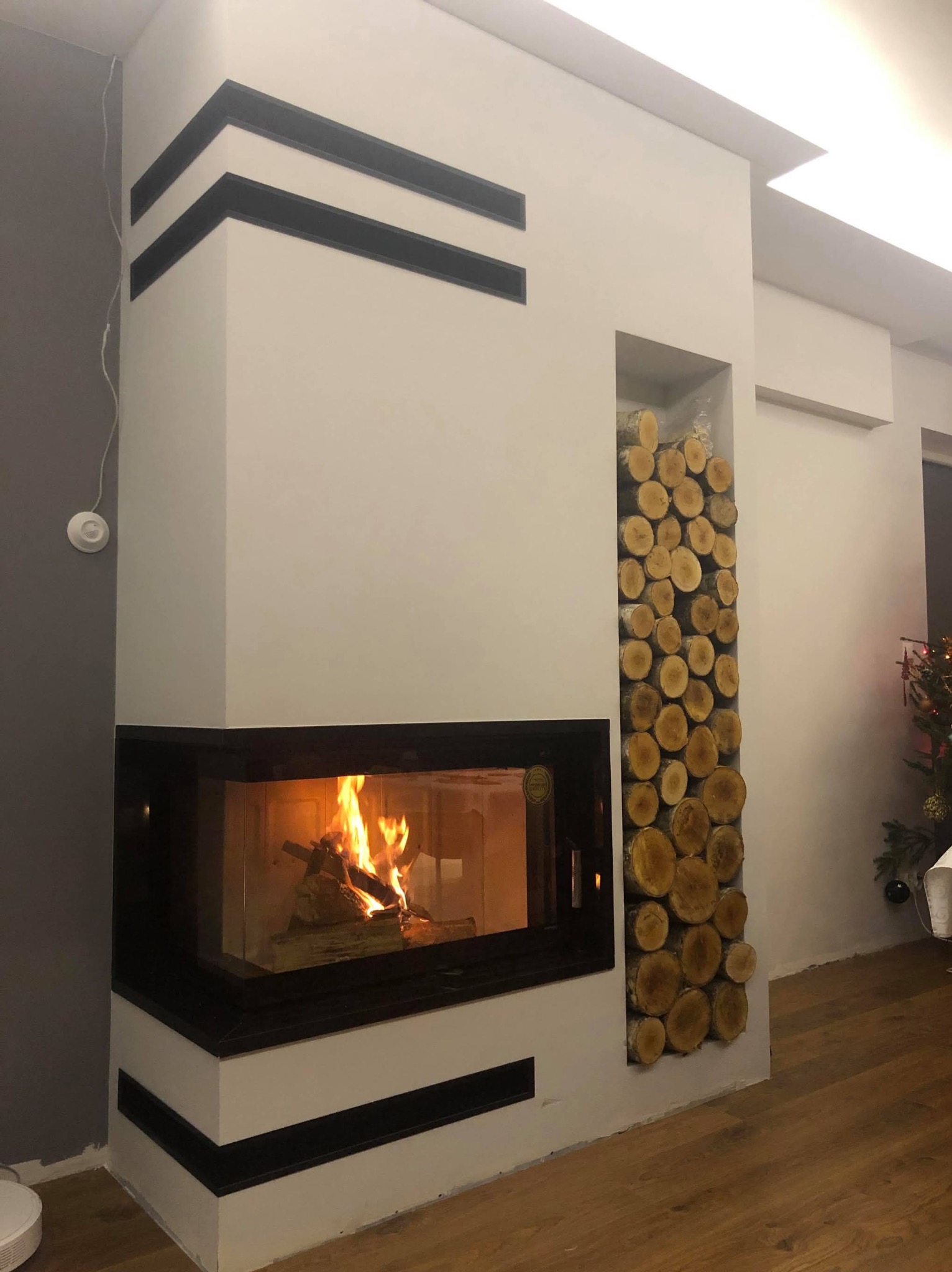 Nowoczesny kominek narożny z szybą, obudowany białą konstrukcją z czarnymi akcentami. Wnęka obok wypełniona dekoracyjnie ułożonymi kawałkami drewna.
