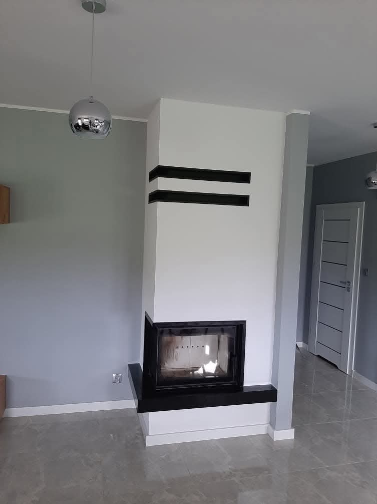 Nowoczesny kominek z czarnymi akcentami na białej obudowie, w szarym wnętrzu z błyszczącą podłogą i srebrną lampą wiszącą.