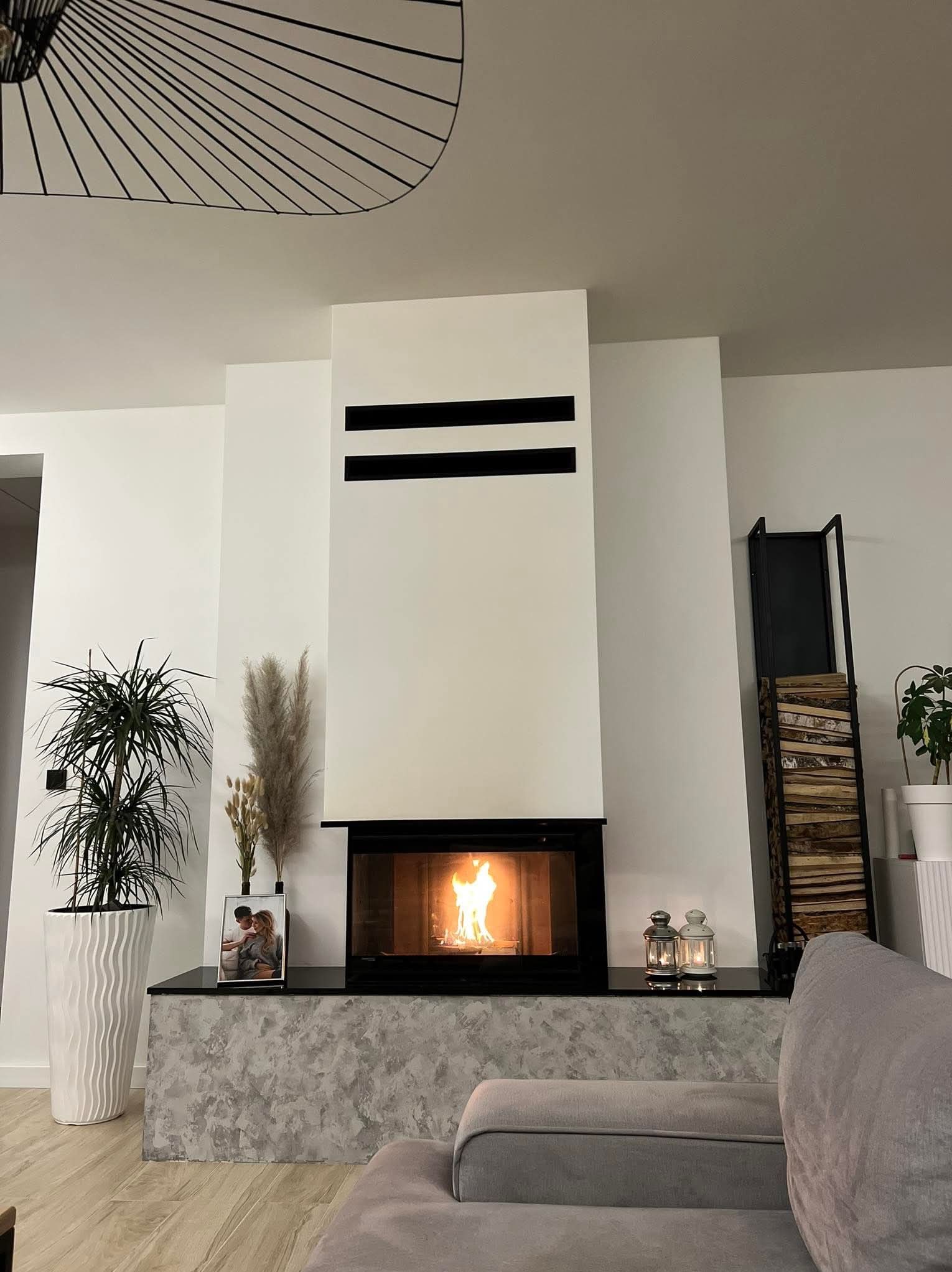 Nowoczesny kominek z czarnymi poziomymi pasami, płomień w palenisku, dekoracje obok. Minimalistyczny design, jasne wnętrze, drewno na półce obok.